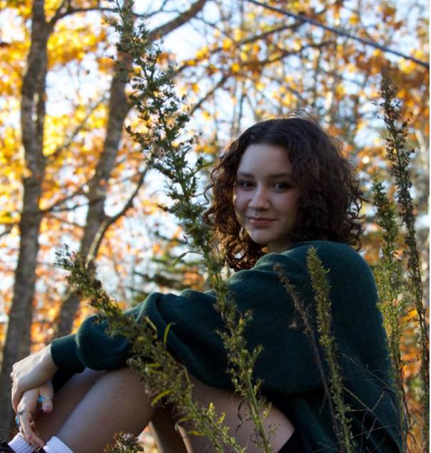 Woman in dark teal sweater sitting among tall green plants with autumn trees in background