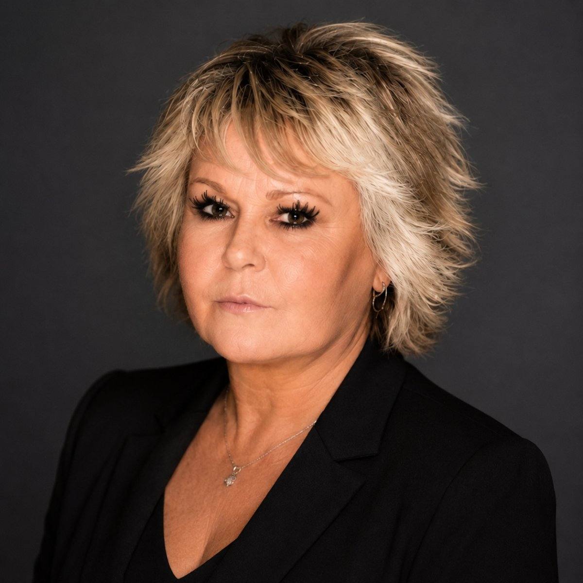 Professional headshot of woman with short blonde layered hair wearing black blazer and silver necklace