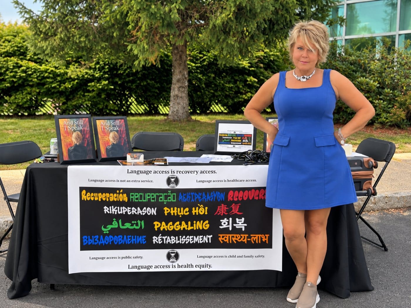 Kim Krawczyk standing beside the And Still I Speak table with framed book covers and a multilingual recovery banner